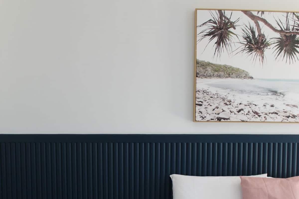 A bedroom with a bed, a bedside table with a lamp, a book, and a wall-mounted painting of a beach scene.