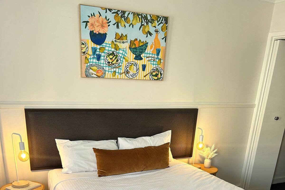 A neatly made bed with a checkered blanket, a headboard, bedside lamps, and a wall art piece above.