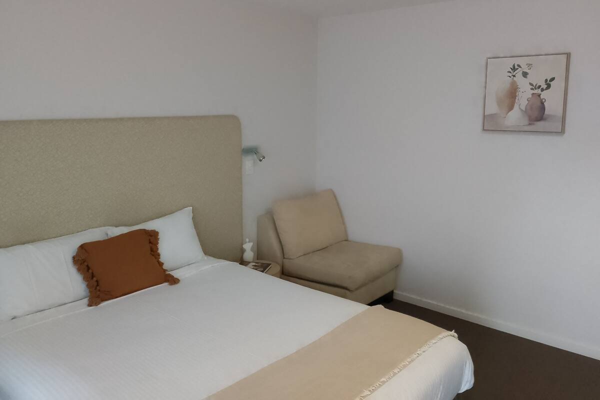 A neatly made bed with white and brown bedding, a beige headboard, and a small armchair in a simple room with white walls and a framed picture on the wall.