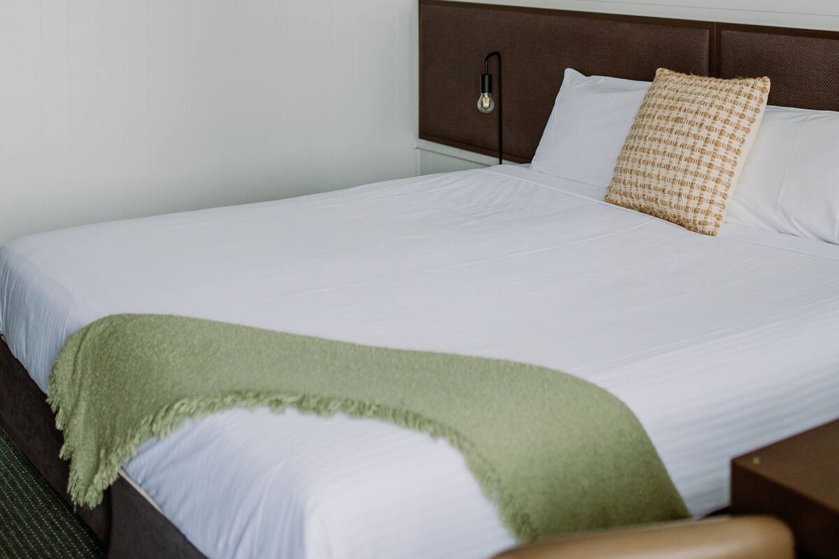 A neatly made bed with white linens, a green throw blanket, and a brown headboard. There are two wooden shelves above the bed, one holding a small plant and a candle, and the other a framed picture.