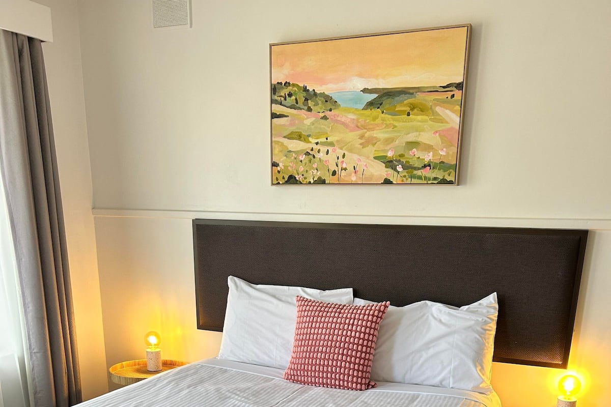 A neatly made bed with white linens and a beige throw blanket, a red pillow, and a headboard with a painting above it.