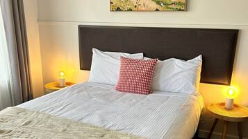 Een net opgemaakt bed met witte lakens, een beige plaid, een rood kussen en een hoofdbord met een schilderij erboven.