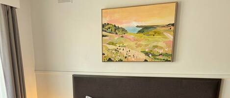 A neatly made bed with white linens and a beige throw blanket, a red pillow, and a headboard with a painting above it.