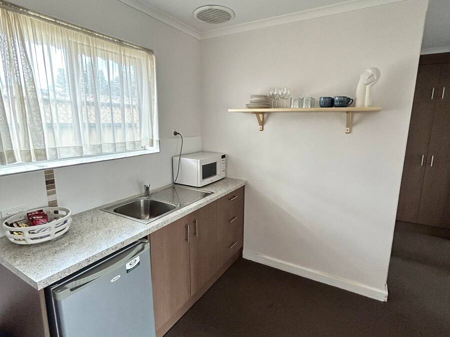A compact kitchen with a dishwasher, a microwave, a sink, and a window with sheer curtains. There is a small shelf with dishes and a white sculpture.