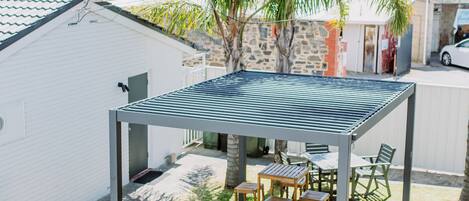A covered patio area with a table and chairs, surrounded by a small lawn and palm trees.