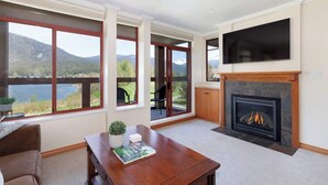 A spacious living room with large windows offering a mountain view, a fireplace, and a flat-screen TV mounted above it. - Green Lake condo w/ panoramic views (Whistler)