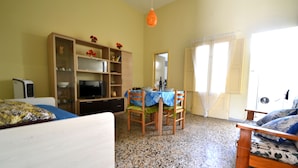 A room with a bed, a TV, a wooden cabinet, a dining table with chairs, and a window with curtains. - Luce Holiday Home With air Conditioning for 5 Guests (Melendugno)