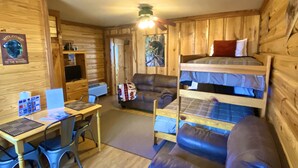 Interior de cabana com decoração em madeira, uma cama beliche, uma escrivaninha com computador, um sofá e uma mesa de jantar.