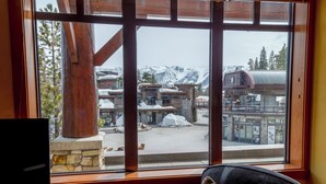 A view from a window with a stone base and wooden beams, overlooking a snow-covered mountain area with buildings and a ski lift.