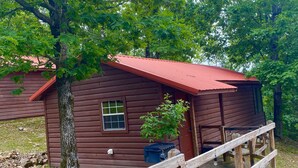 A rustic cabin with a red roof, surrounded by trees. A wooden deck leads to the cabin, and a sign reads "FISHERMANS COVE." - Fisherman's Cove (Mena)