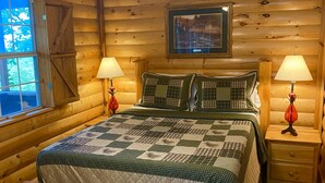 A wooden cabin bedroom with a bed, two bedside tables with lamps, a ceiling fan, and a window with a view of trees. - Fisherman's Cove (Mena)