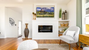 Un salon moderne avec un téléviseur à écran plat fixé au-dessus d’une cheminée, un plancher en bois, un fauteuil blanc et une table basse.