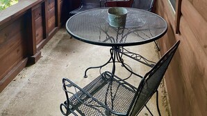 Ein überdachter Außenbereich mit einem runden Metalltisch, vier Stühlen, einem Metal-Eimer und einer Bank entlang des Holzgeländers.