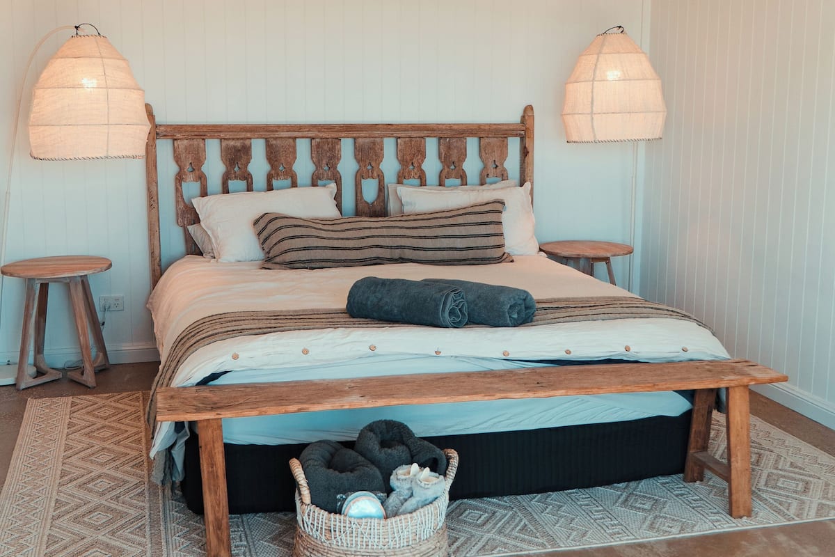 A bedroom with a wooden bed, a ceiling fan, two bedside lamps, a small round table, and a woven basket on the floor.