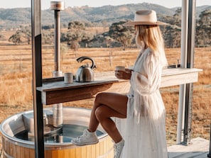 Una persona sentada junto a una mesa de madera, con un hervidor y una taza, con vistas a un campo seco y colinas al fondo.