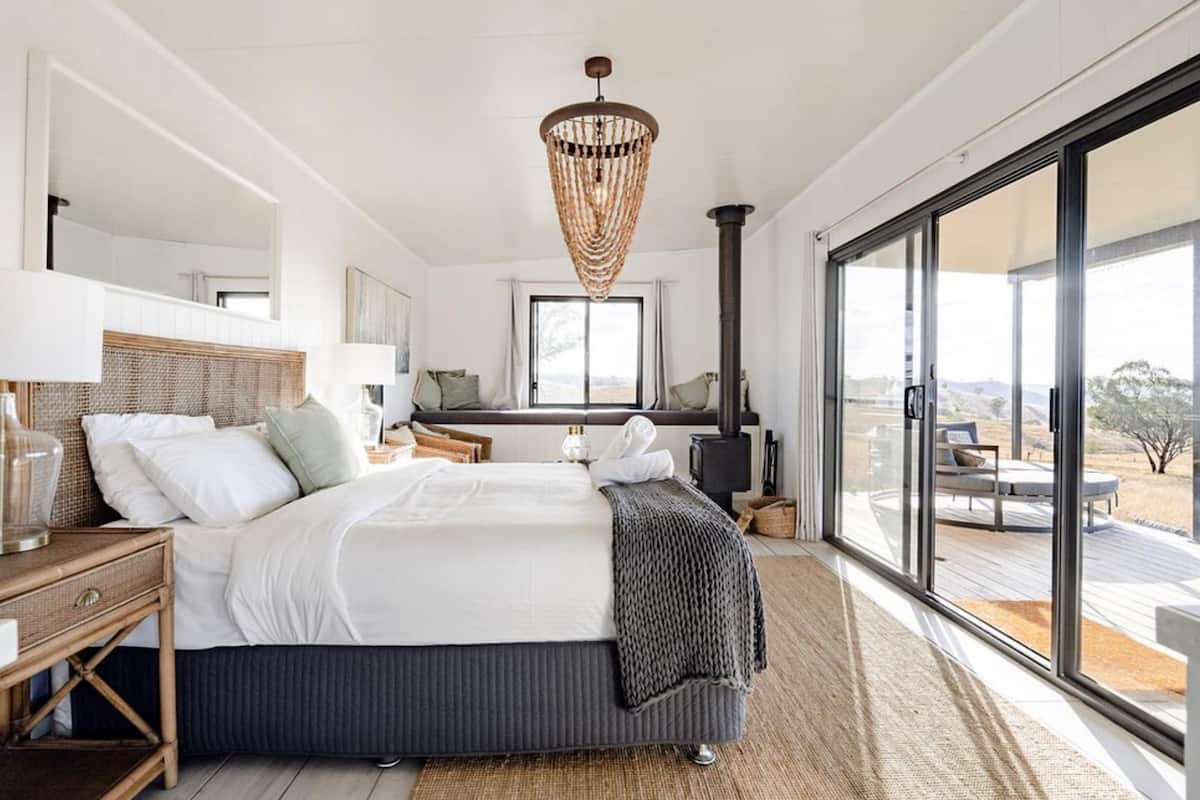 A modern bedroom with a large bed, a bedside table, a lamp, a woven ceiling light, a view of the outdoors through large windows, and a fireplace.