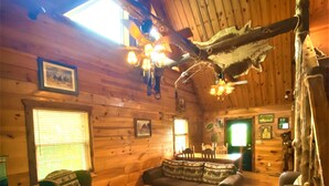 Interior acogedor de una cabaña de troncos con paredes y techo de madera, y un ventanal grande. Hay varios muebles, incluyendo un sofá, sillas y una mesa de centro.