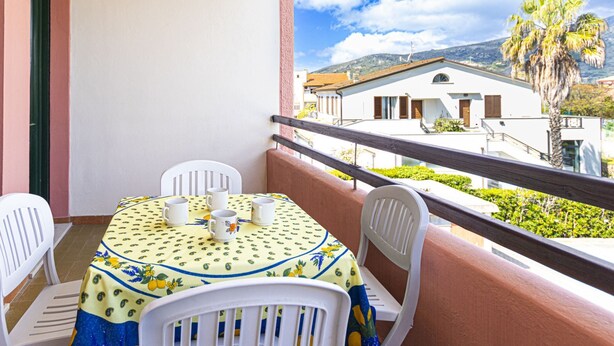A balcony with a table set for two, chairs, and a view of a building and palm trees. - Carlotta, apartment a few steps from the pedestrian center of Marina di Campo equipped with air cond (Marina di Campo)