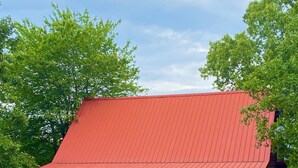 A rustic log cabin with a red roof, a porch, and a gravel ground.