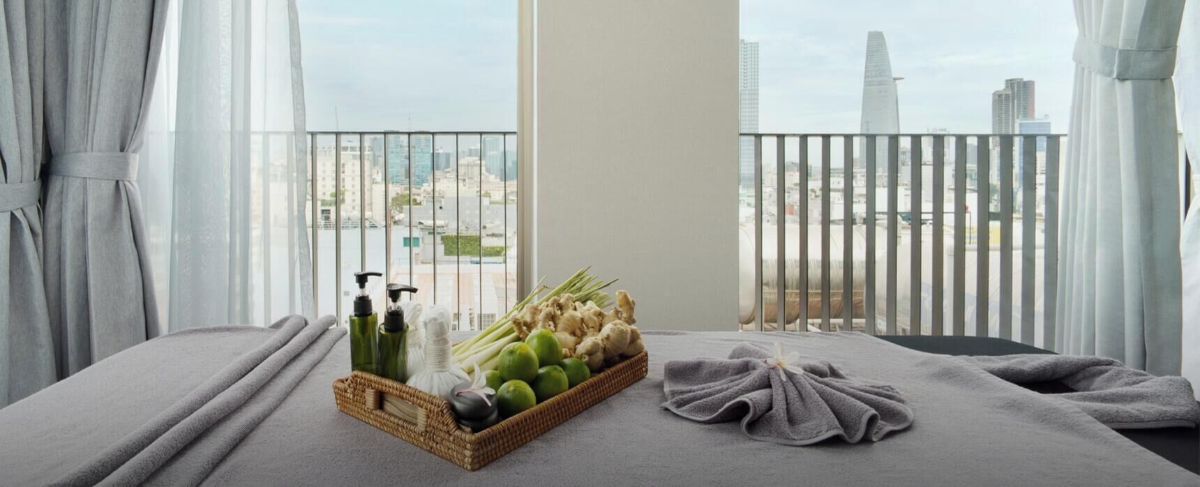Un cuarto de spa con una camilla de masaje, una cesta de hierbas frescas y vistas de la ciudad a través de la barandilla de un balcón.