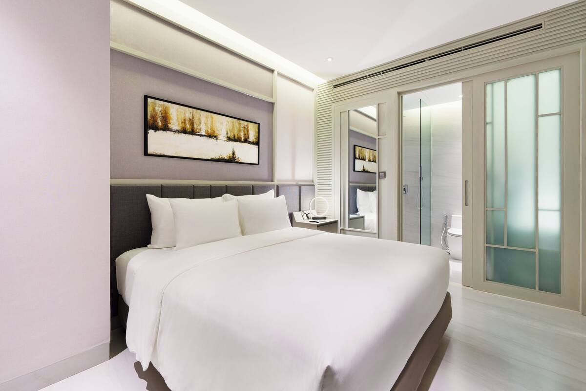 Un dormitorio de hotel moderno con una cama grande, una mesita de noche, un cuadro en la pared y un cerramiento de vidrio corredizo que da a un baño.