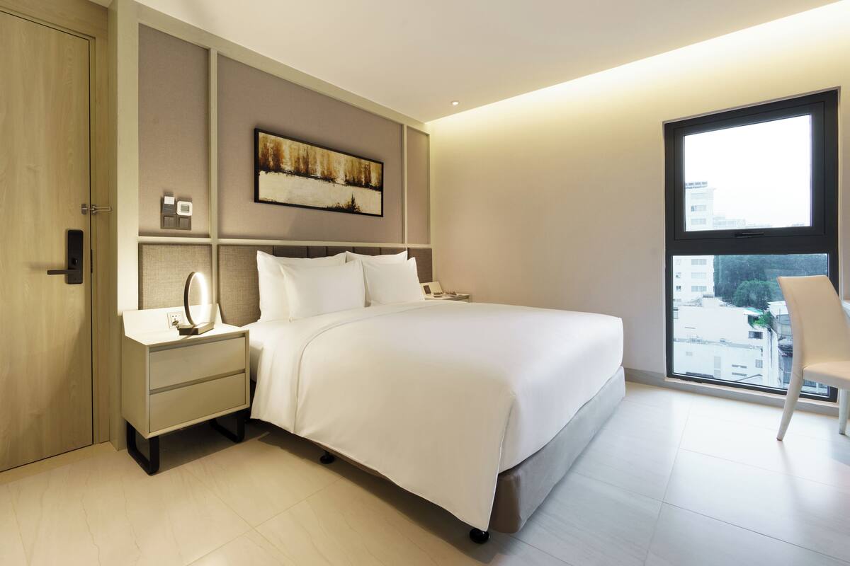 Un dormitorio moderno con una cama grande, una mesita de noche con espejo, una lámpara auxiliar y un cuadro colgado en la pared, además de un ventanal con vistas a edificios.