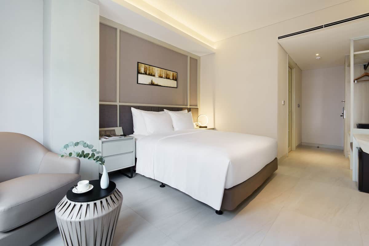 Una habitación de hotel moderna con una cama grande, una mesita de noche con lámpara, una silla y una pequeña mesa redonda con un florero.