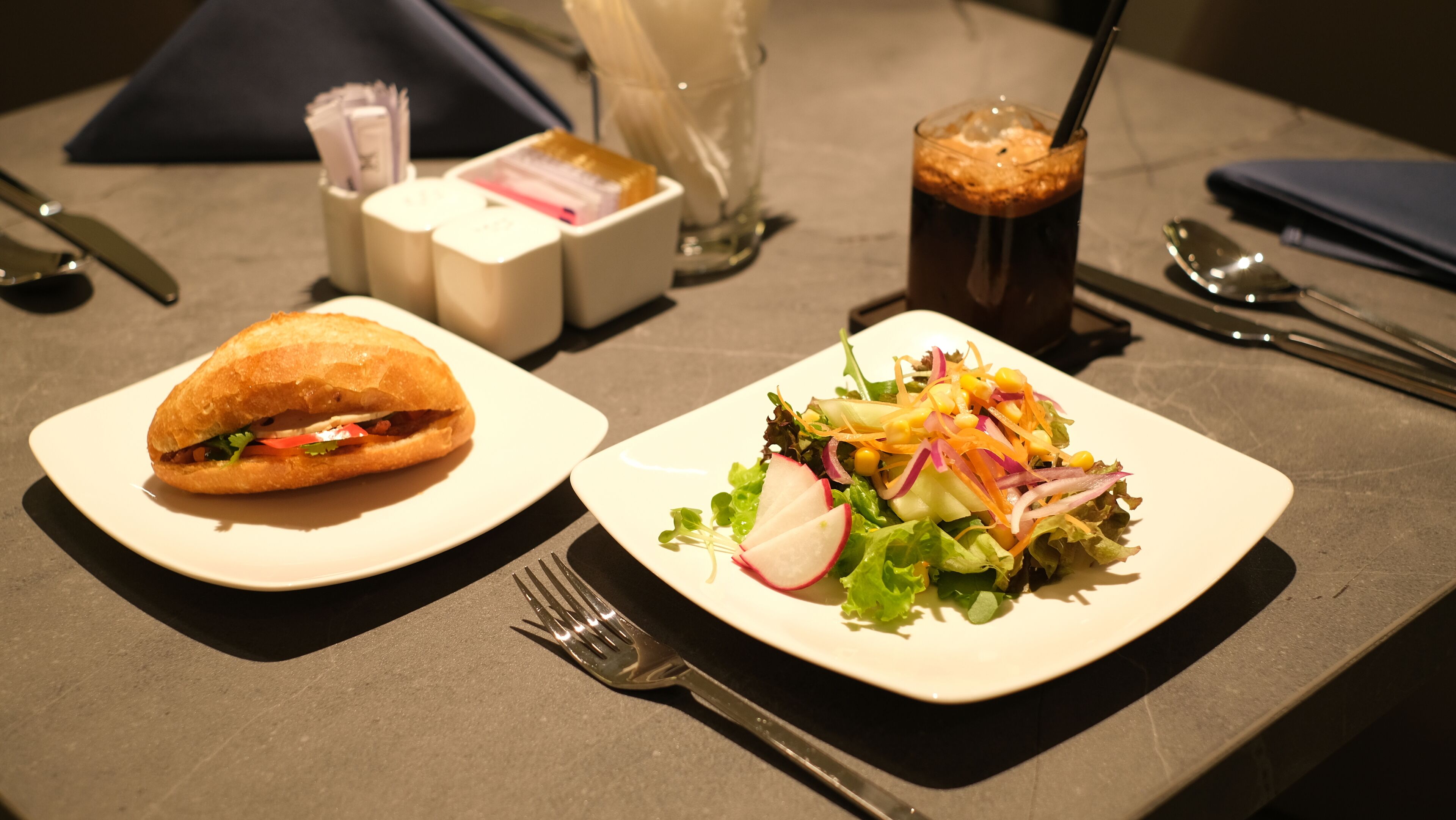 A table with a sandwich and a salad on white plates, a drink, and cutlery.