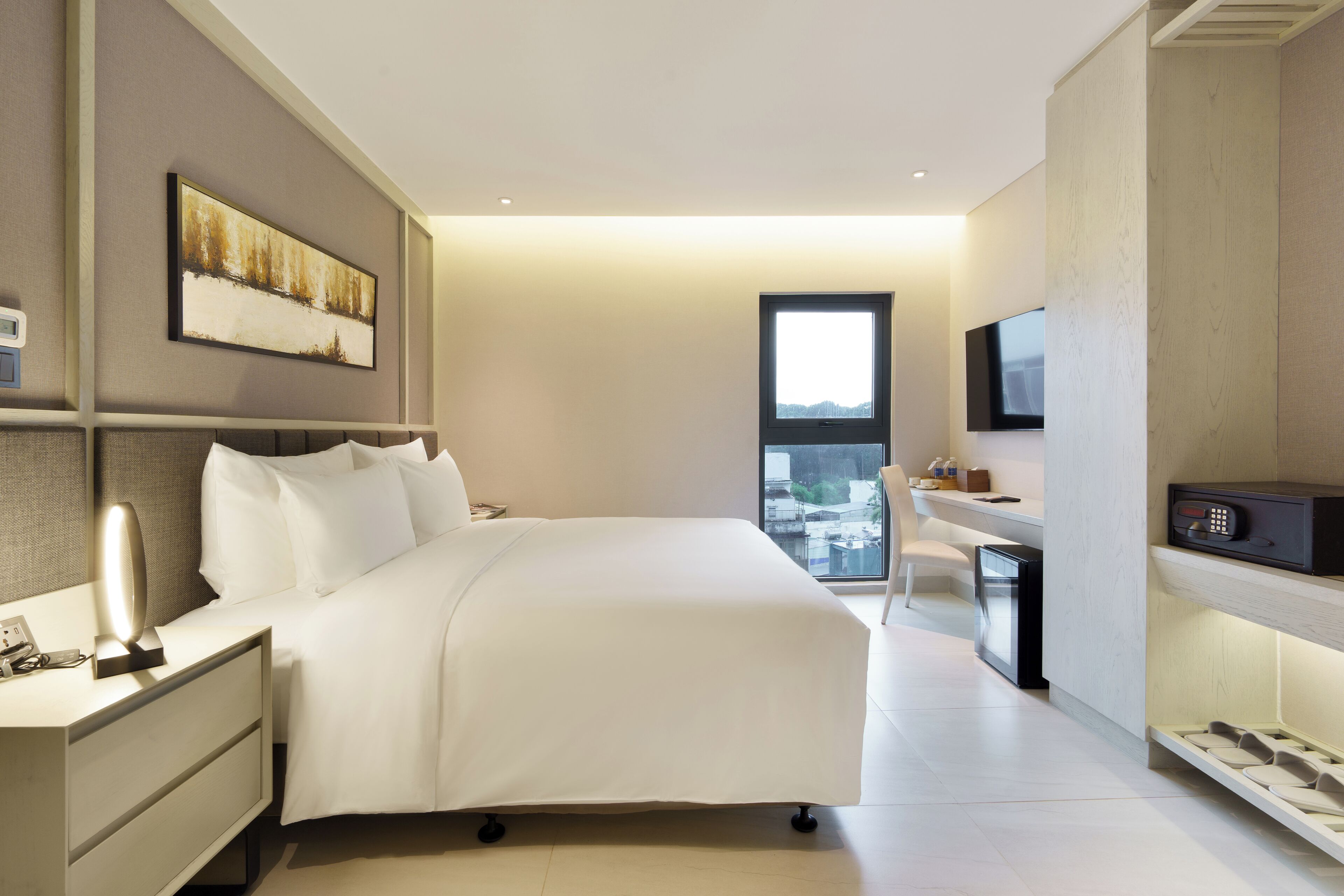 A modern hotel room with a large bed, bedside table with a lamp, a desk with a chair, a TV mounted on the wall, and a window offering a city view.