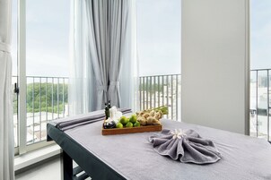 A massage table with a towel, a tray of fruits, and a bottle of oil in a hotel room with a balcony view.