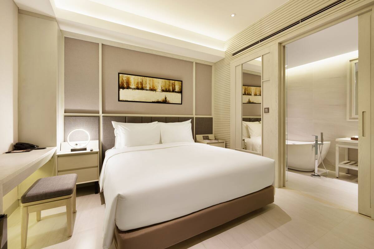 Una habitación de hotel moderna con una cama grande, mesitas de noche, un escritorio con silla, un espejo, un cuadro en la pared y un baño con bañera y ducha.