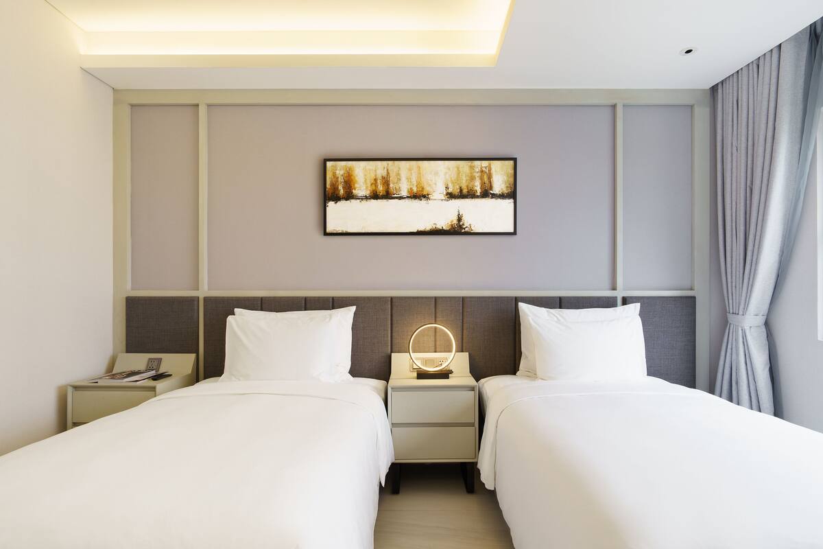 Habitación de hotel con dos camas, una mesita de noche con lámpara, un cuadro en la pared y una ventana con cortinas.