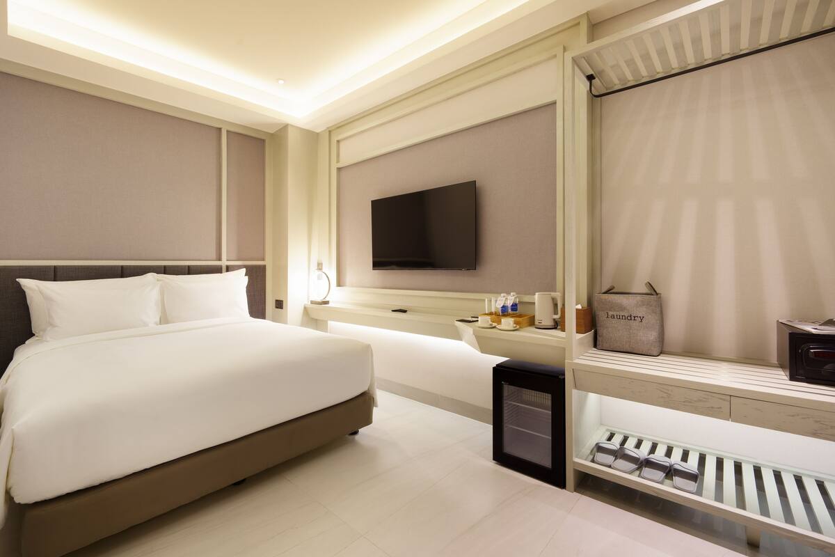 Una habitación de hotel moderna con una cama grande, un televisor de pantalla plana en la pared, una mesita de noche con lámpara y un estante pequeño con vasos y un cesto de ropa.