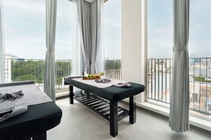 A modern room with a massage table, a small table with towels and fruits, large windows with curtains, and a city view.