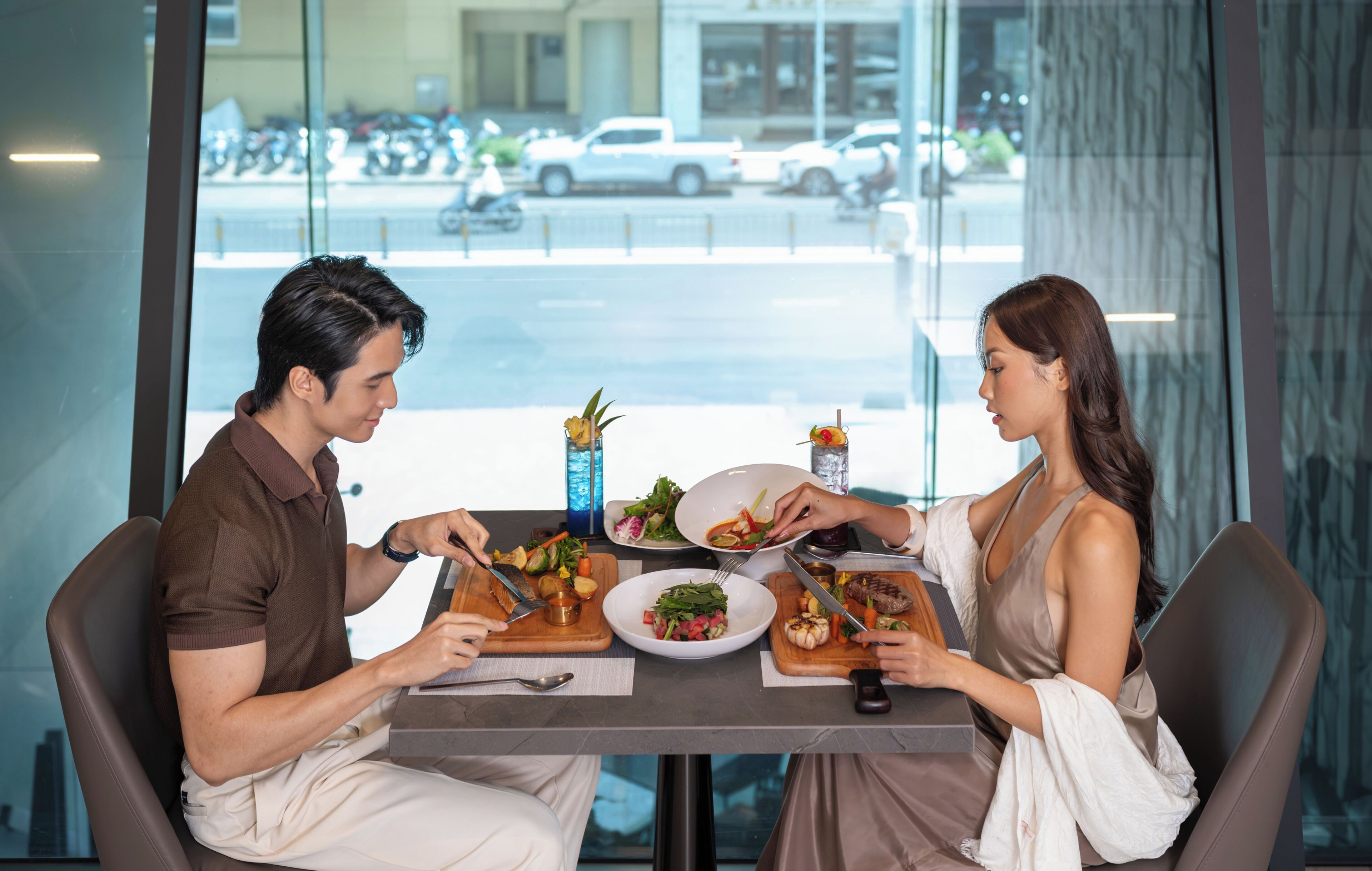 A dining table with food, a man and a woman eating, and a view of the street outside.