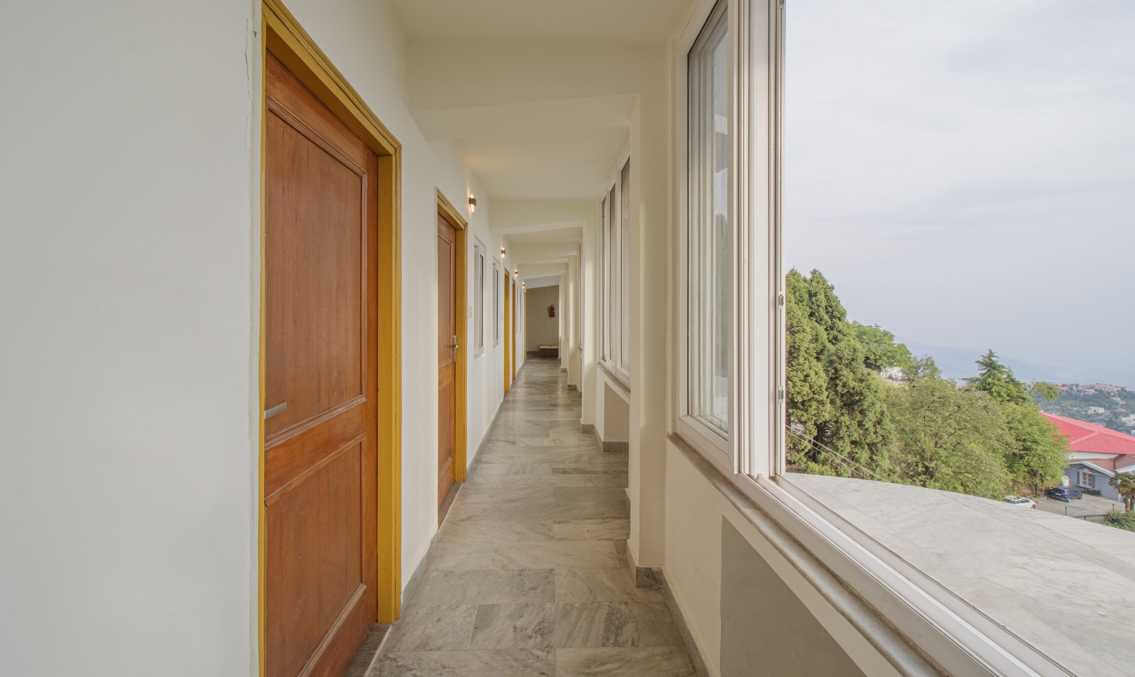 A corridor with wooden doors and windows, leading to a room with a view of trees and buildings.