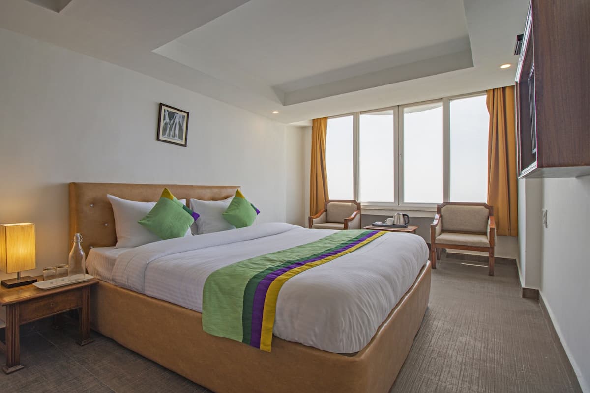 A hotel room with a large bed, a bedside table with a lamp, a chair, and a window with curtains.
