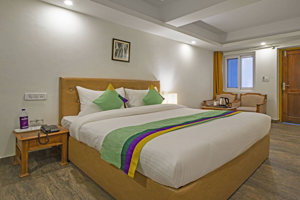 A hotel room with a large bed, a wooden headboard, a nightstand with a phone and a bottle, a chair, and a wooden door.