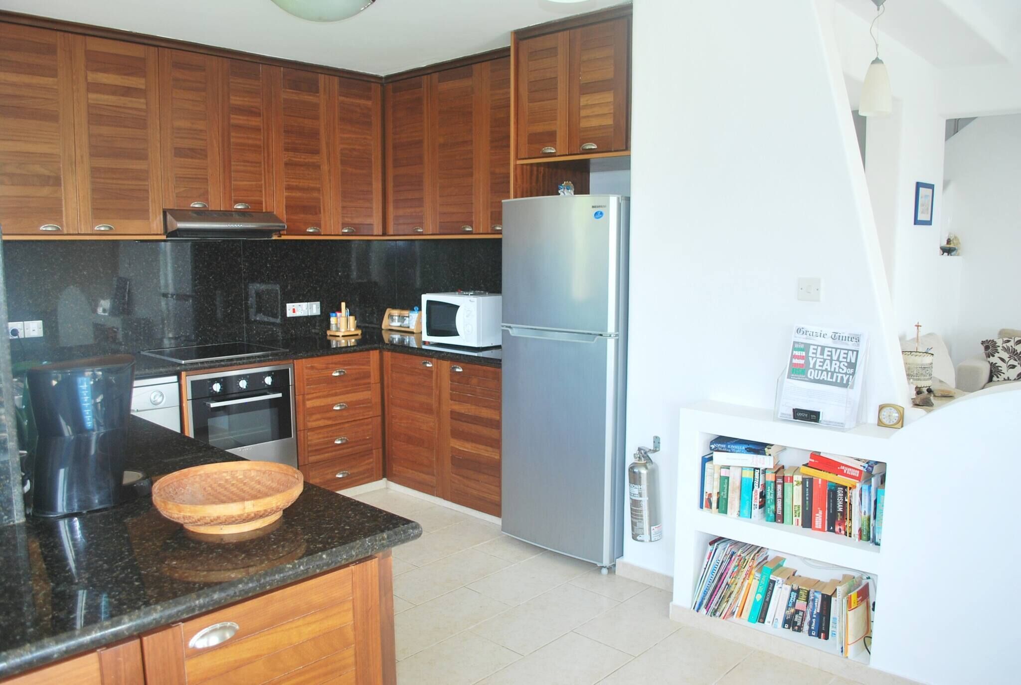 A kitchen with wooden cabinets, a microwave, a refrigerator, a coffee maker, and a bookshelf with books.