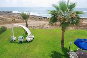 A grassy area with a boat, chairs, and a table, overlooking a rocky beach and ocean. - Sea Front Villa With Private Heated Pool, Quiet Area Paphos 322 (Kissonerga)