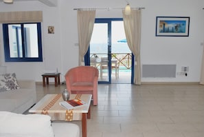 Une pièce avec un canapé, une table sur laquelle se trouve un magazine, un fauteuil rose, une fenêtre avec des rideaux et une vue sur la mer.