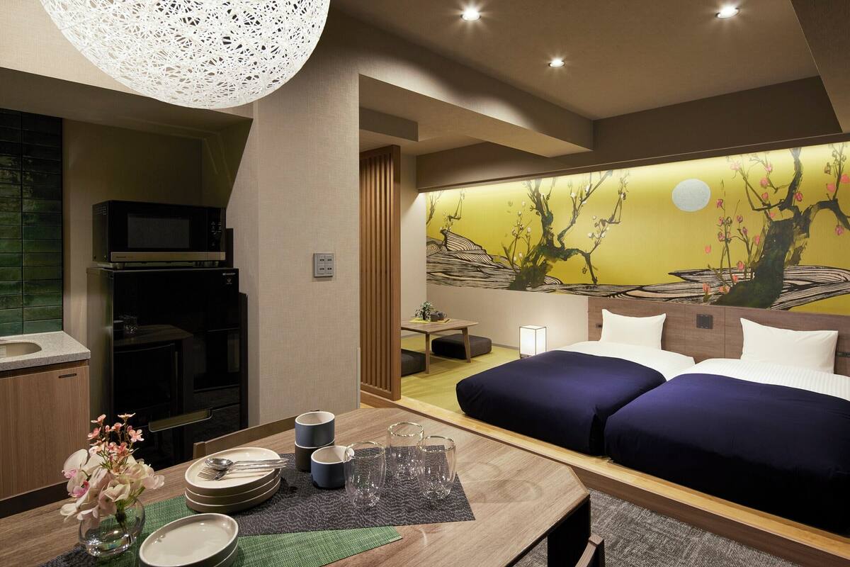 A modern hotel room with a dining area, a bed with dark blue bedding, a wall mural, and a kitchenette.