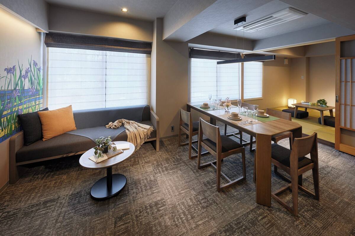 A modern hotel room with a sofa, a dining table, and chairs. There is a window with blinds, a painting on the wall, and a carpeted floor.