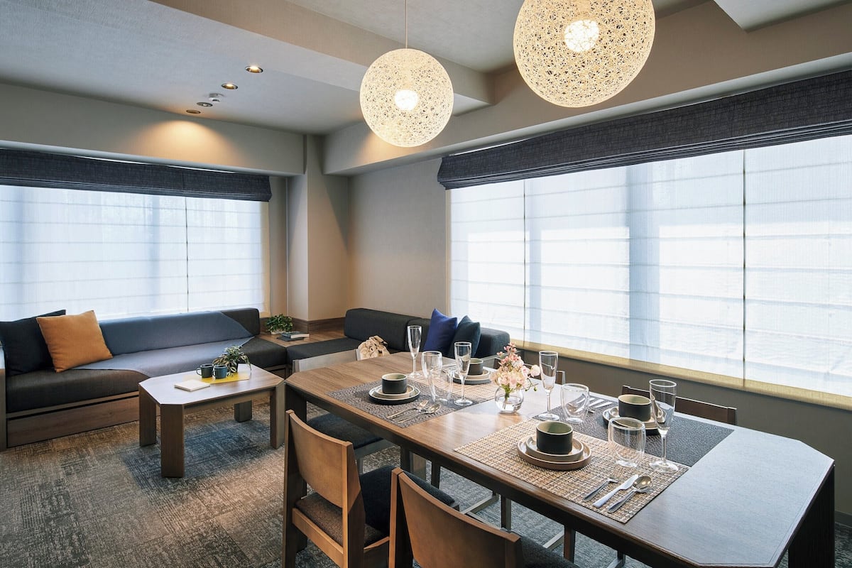 A modern hotel room with a dining area, a sofa, and large windows.