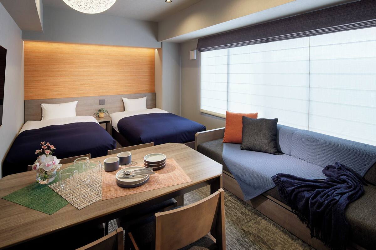 A hotel room with two beds, a sofa, a dining table with plates and glasses, and a large window with blinds.
