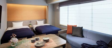 A hotel room with two beds, a sofa, a dining table with plates and glasses, and a large window with blinds.
