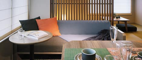 Une pièce de style japonais moderne, avec un canapé, une petite table ronde sur laquelle se trouvent un livre et des lunettes, et un coin repas agrémenté d’un tapis vert, d’assiettes et de couverts.