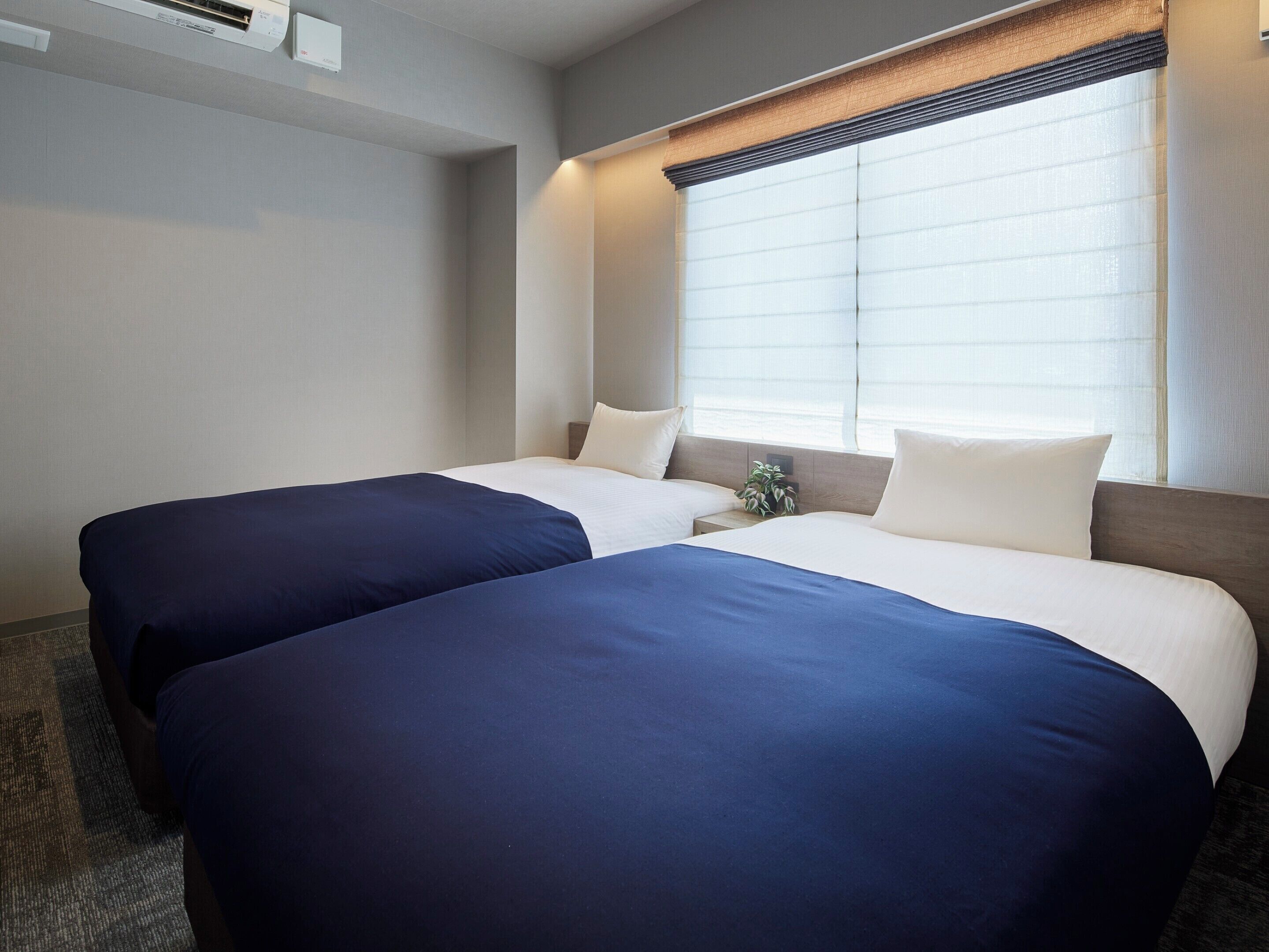 Una habitación de hotel con dos camas, una ventana con persianas, una mesita de noche con una planta y un aire acondicionado de pared.