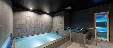A modern indoor swimming pool with a geometric wall design, a glass-enclosed sauna, and a tiled floor.