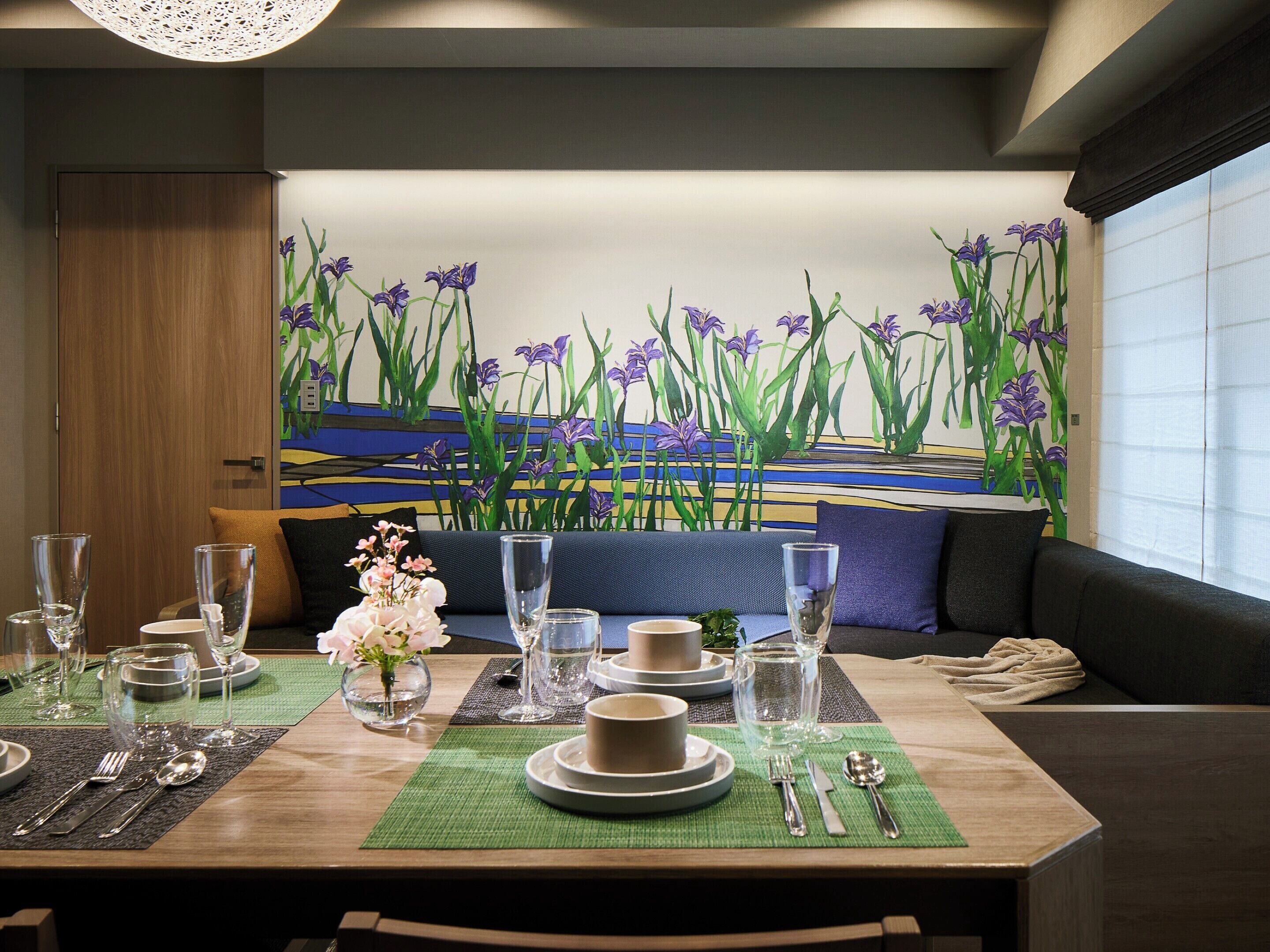 Un comedor con una mesa dispuesta para dos personas, con un cuadro de flores moradas en la pared de fondo.